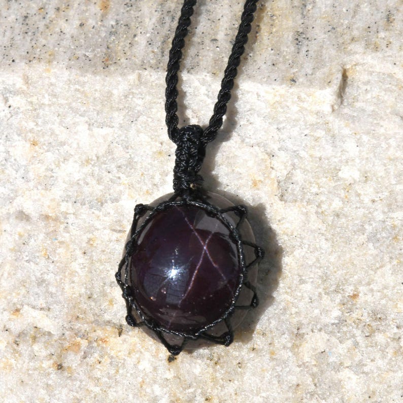 Close-up of a polished star garnet pendant in deep burgundy, wine-red, and dark plum tones, displayed on a light stone background. A distinct four-rayed star appears across the gemstone when illuminated, creating a bright reflective cross. The garnet has a smooth glossy finish with rich color depth and natural translucency. It is secured in a black macrame net and attached to a black adjustable cord. Associated with passion, vitality, grounding, courage, and emotional strength.