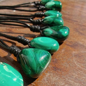 Close-up of multiple polished irregular Malachite in shades of vivid green and deep forest green, arranged in a row on a wooden surface. The green opaque gemstones have smooth glossy surfaces with distinct banded patterns, concentric rings, and flowing lines, with light reflections highlighting their curved shapes. It is attached to a black cord with a top knot and adjustable knot closure. Malachite is associated with transformation, protection, and emotional healing.