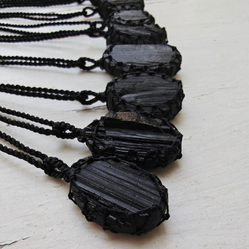 Close-up of multiple raw oval Black Tourmaline in deep black shades, resting on a white wood surface. The Black Tourmaline dark gemstone has strong parallel vertical striations, rough uneven edges, and a matte to slightly reflective texture with natural ridges and occasional lighter inclusions and surface variations. Each piece is wrapped in a black macrame net and attached to a black cord with an adjustable knot closure. Black Tourmaline is associated with grounding and protection.