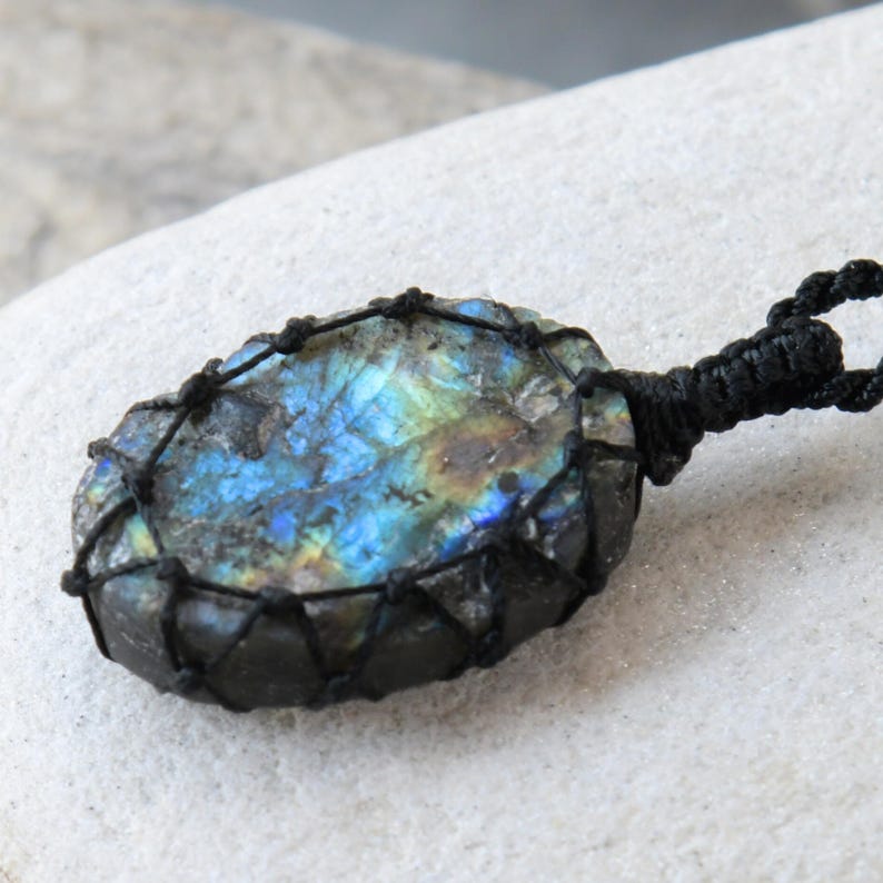 Raw Labradorite pendant displaying vivid flashes of electric blue, turquoise, gold, and hints of green across its natural rough surface. The stone is wrapped in a black macrame net with an adjustable black cord with a sliding knot closure and crystal beads and displayed on a light stone background. Labradorite is associated with transformation, intuition, protection, inner strength, spiritual awakening,  and enhanced creativity.
