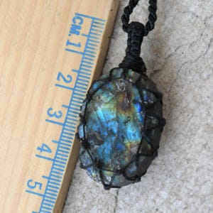Raw Labradorite pendant displaying vivid flashes of electric blue, turquoise, gold, and hints of green across its natural rough surface. The stone is wrapped in a black macrame net with an adjustable black cord with a sliding knot closure and crystal beads and displayed on a light stone background beside a wooden ruler. Labradorite is associated with transformation, intuition, protection, inner strength, spiritual awakening,  and enhanced creativity.