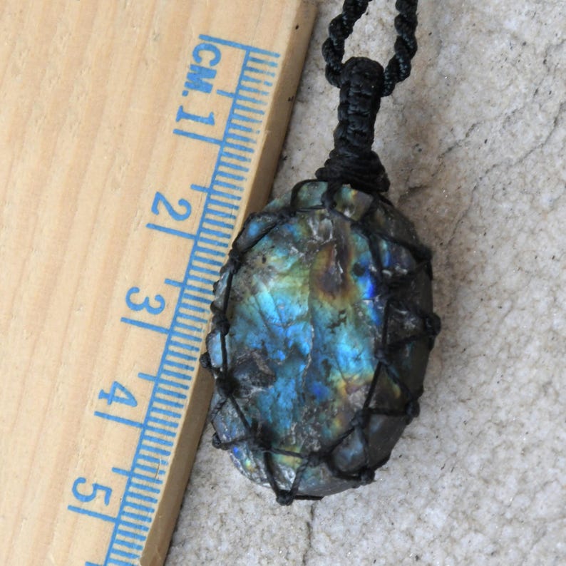 Raw Labradorite pendant displaying vivid flashes of electric blue, turquoise, gold, and hints of green across its natural rough surface. The stone is wrapped in a black macrame net with an adjustable black cord with a sliding knot closure and crystal beads and displayed on a light stone background beside a wooden ruler. Labradorite is associated with transformation, intuition, protection, inner strength, spiritual awakening,  and enhanced creativity.