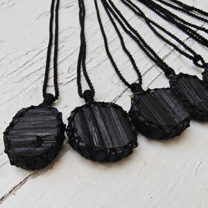 Close-up of multiple raw oval and round Black Tourmaline in deep black shades, resting on a white wood surface. The Black Tourmaline dark gemstone has strong parallel vertical striations, rough uneven edges, and a matte to slightly reflective texture with natural ridges and occasional lighter inclusions and surface variations. Each piece is wrapped in a black macrame net and attached to a black cord with an adjustable knot closure. Black Tourmaline is associated with grounding and protection.