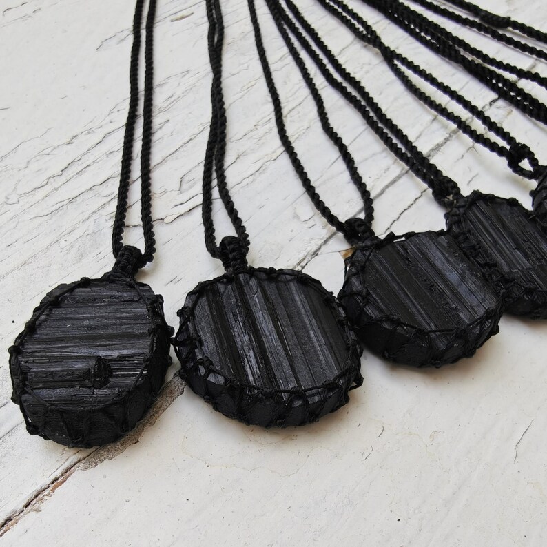 Close-up of multiple raw oval and round Black Tourmaline in deep black shades, resting on a white wood surface. The Black Tourmaline dark gemstone has strong parallel vertical striations, rough uneven edges, and a matte to slightly reflective texture with natural ridges and occasional lighter inclusions and surface variations. Each piece is wrapped in a black macrame net and attached to a black cord with an adjustable knot closure. Black Tourmaline is associated with grounding and protection.