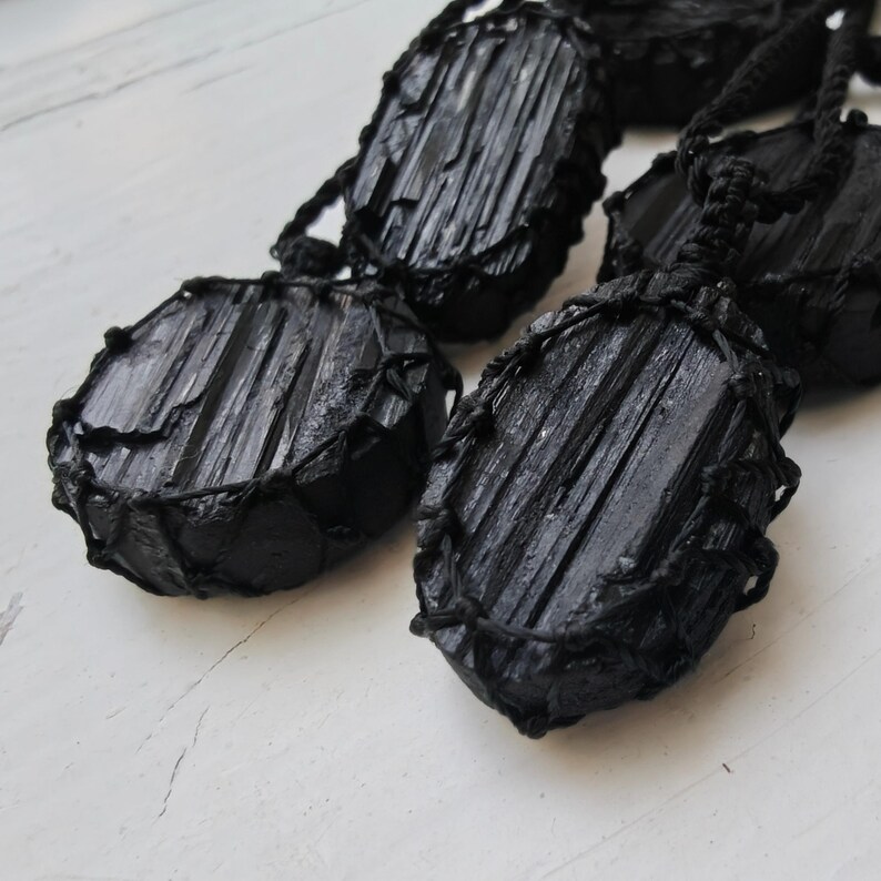 Close-up of multiple raw oval and round Black Tourmaline in deep black shades, resting on a white wood surface. The Black Tourmaline dark gemstone has strong parallel vertical striations, rough uneven edges, and a matte to slightly reflective texture with natural ridges and occasional lighter inclusions and surface variations. Each piece is wrapped in a black macrame net and attached to a black cord with an adjustable knot closure. Black Tourmaline is associated with grounding and protection.