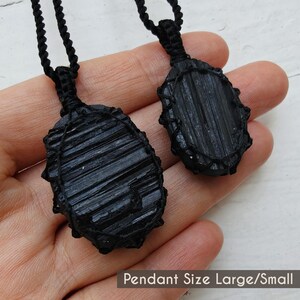 Close-up of 2 raw oval Black Tourmaline in deep black shades, displayed on a hand against a white background. The Black Tourmaline dark gemstone has strong parallel vertical striations, rough uneven edges, and a matte to slightly reflective texture with natural ridges and minor surface fractures. Each piece is wrapped in a black macrame net and attached to a black cord with an adjustable knot closure. The image includes the text “Pendant Size Large/Small”.