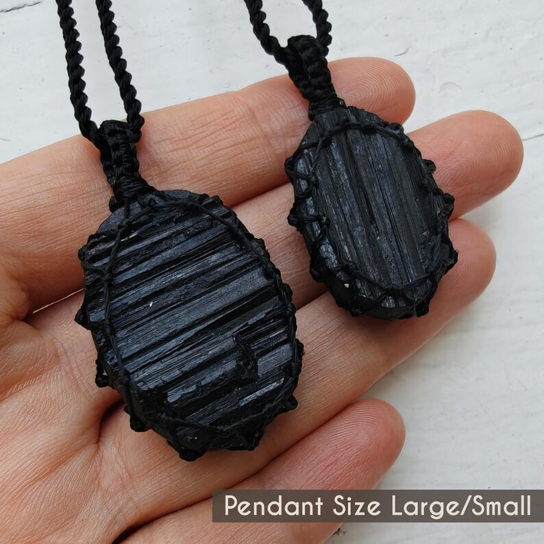 Close-up of 2 raw oval Black Tourmaline in deep black shades, displayed on a hand against a white background. The Black Tourmaline dark gemstone has strong parallel vertical striations, rough uneven edges, and a matte to slightly reflective texture with natural ridges and minor surface fractures. Each piece is wrapped in a black macrame net and attached to a black cord with an adjustable knot closure. The image includes the text “Pendant Size Large/Small”.