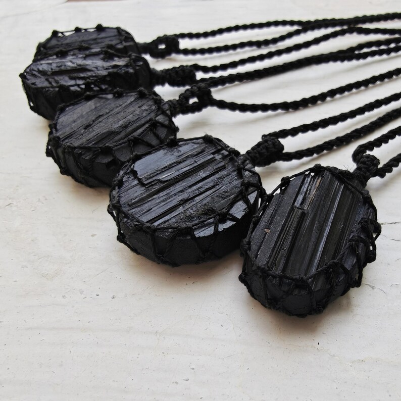 Close-up of 5 raw oval and round Black Tourmaline in deep black shades, resting on a white wood surface. The Black Tourmaline dark gemstone has strong parallel vertical striations, rough uneven edges, and a matte to slightly reflective texture with natural ridges and occasional lighter inclusions and surface variations. Each piece is wrapped in a black macrame net and attached to a black cord with an adjustable knot closure. Black Tourmaline is associated with grounding and protection.