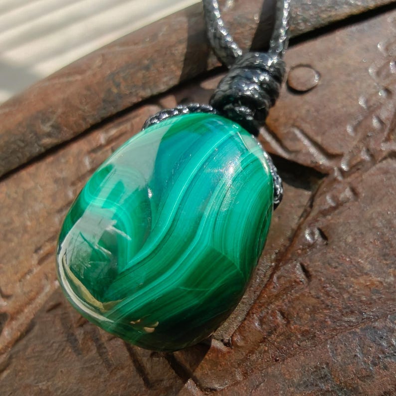 Close-up of a polished teardrop Malachite in shades of vivid green and deep forest green, resting on a textured brown surface. The green opaque gemstone has a smooth glossy surface with distinct banded patterns, flowing concentric lines, and layered swirls, with light reflections highlighting its curved form. It is attached to a black cord with a top knot and adjustable knot closure. Malachite is associated with transformation, protection, and emotional healing.
