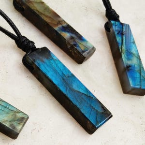 Op de afbeelding: Drie rechthoekige hangerkettingen met gepolijste labradorietstenen met iriserende blauwe en gouden flitsen. Elke steen hangt aan een zwart koord met een geknoopte bevestiging, met natuurlijke minerale texturen en diepblauwe tinten.