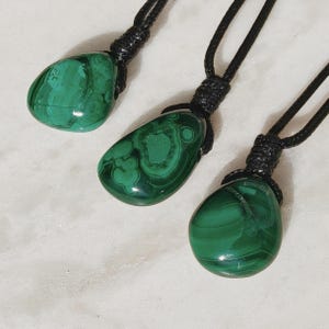 Close-up of 3 polished irregular Malachite in shades of vivid green and deep forest green, arranged on a light stone surface. The green opaque gemstones have smooth glossy surfaces with distinct banded patterns, concentric lines, and varied formations, including rounded and triangular shapes with layered green stripes and darker swirls. It is attached to a black cord with a top knot and adjustable knot closure. Malachite is associated with transformation, protection, and emotional healing.