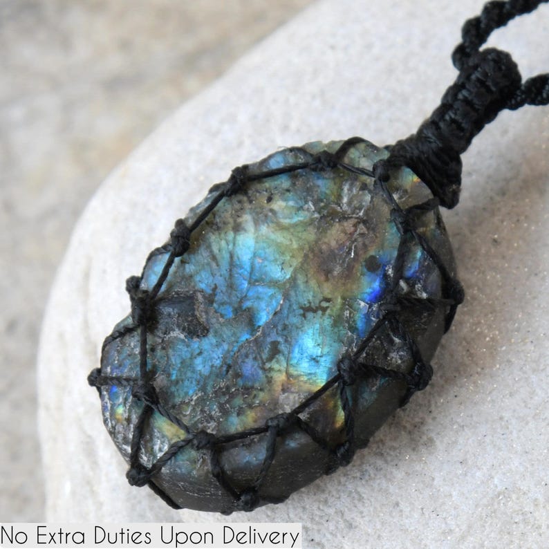 A single, raw, druzy oval Labradorite necklace pendant is displayed on a textured light gray rock surface. The gemstone exhibits strong flashes of blue, green, and golden-brown (labradorescence). It is encased in a black macrame net that forms the necklace cord with a sliding knot closure and crystal beads. Labradorite is associated with intuition, spiritual awareness, protection, and transformation.