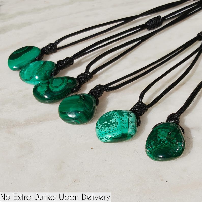 Close-up of 6 polished irregular Malachite in shades of vivid green and deep forest green, arranged on a light stone surface. The green opaque gemstones have smooth glossy surfaces with distinct banded patterns, concentric lines, and varied formations, including rounded and triangular shapes with layered green stripes and darker swirls. It is attached to a black cord with a top knot and adjustable knot closure. Malachite is associated with transformation, protection, and emotional healing.