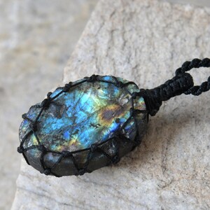 Raw Labradorite pendant displaying vivid flashes of electric blue, turquoise, gold, and hints of green across its natural rough surface. The stone is wrapped in a black macrame net with an adjustable black cord with a sliding knot closure and crystal beads and displayed on a light stone background. Labradorite is associated with transformation, intuition, protection, inner strength, spiritual awakening,  and enhanced creativity.