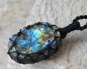 Puede incluir: Un collar con colgante de labradorita. La piedra de forma ovalada está envuelta en un cordón negro tejido, con un lazo de cordón negro para llevarlo. La piedra tiene un brillo iridiscente azul, verde y amarillo. El collar se muestra sobre una superficie de piedra gris claro.