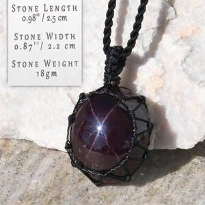 Polished star garnet pendant in deep burgundy, dark red- plum tones, displayed on a white background. A distinct four-rayed star appears across the gemstone when illuminated by direct light, creating a bright reflective cross. Text on the image states: Stone Length 0.98" / 2.5 cm, Stone Width 0.87" / 2.2 cm, Stone Weight 18 gm. The pendant is secured in a black macrame net and attached to a black adjustable cord. Associated with passion, vitality, grounding, courage, and emotional strength.