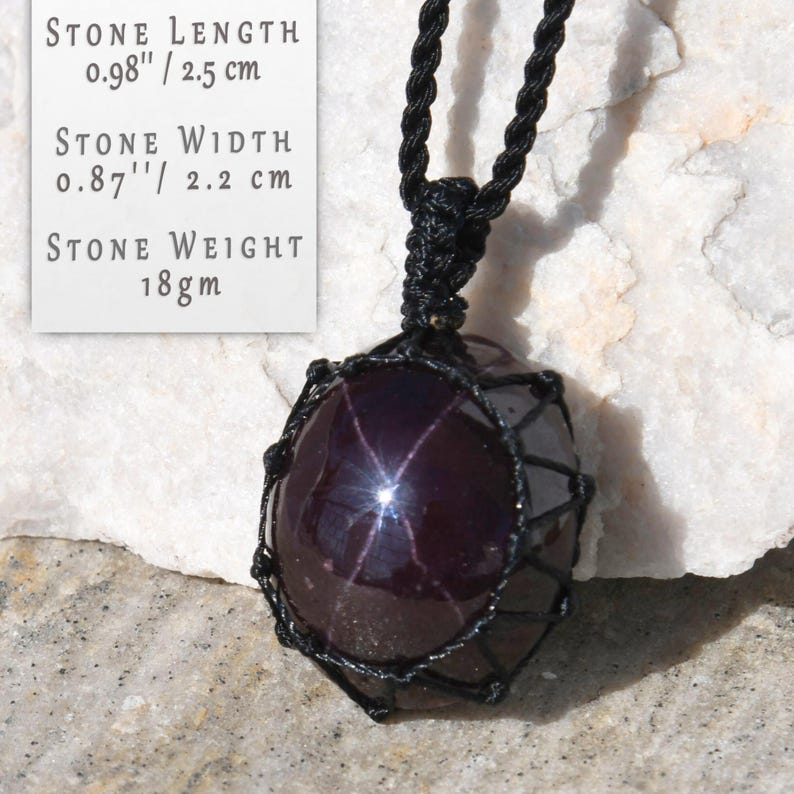 Polished star garnet pendant in deep burgundy, dark red- plum tones, displayed on a white background. A distinct four-rayed star appears across the gemstone when illuminated by direct light, creating a bright reflective cross. Text on the image states: Stone Length 0.98" / 2.5 cm, Stone Width 0.87" / 2.2 cm, Stone Weight 18 gm. The pendant is secured in a black macrame net and attached to a black adjustable cord. Associated with passion, vitality, grounding, courage, and emotional strength.