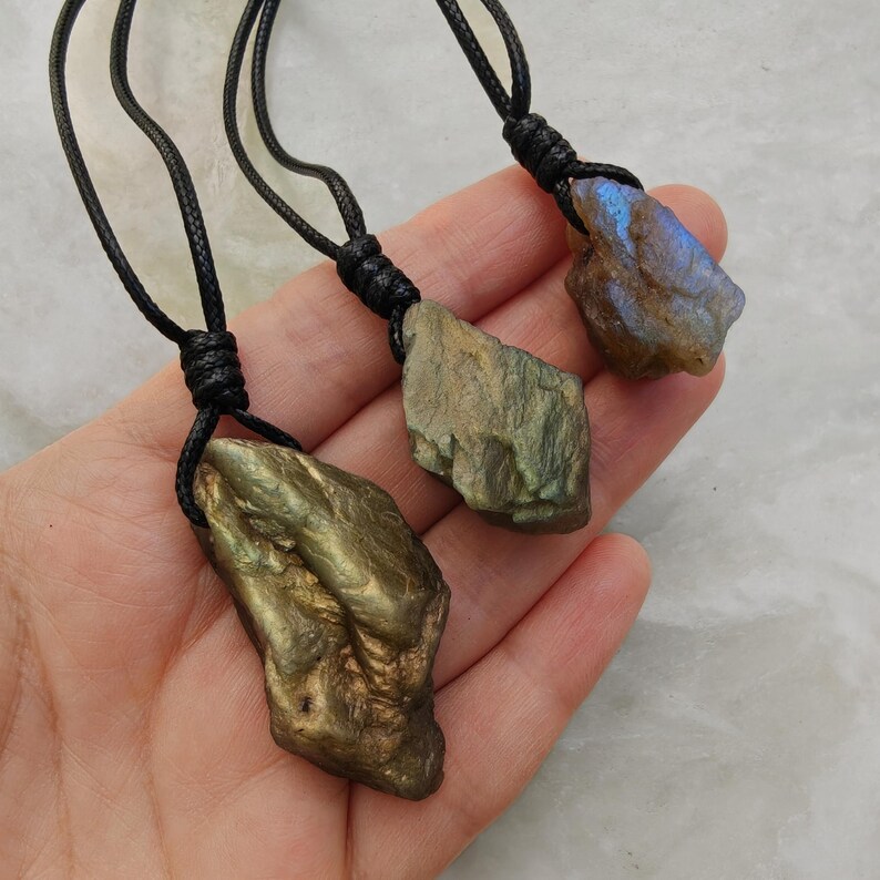 Close-up of three raw irregular Labradorite pendants in shades of gold, green, and blue, displayed on a hand. The Labradorite gemstones have a rough natural texture with strong iridescent flashes, layered color play, and visible surface striations. Each stone is secured with a top knot and attached to a black cord with an adjustable knot closure. Labradorite is associated with transformation, protection, intuition, and spiritual shielding.