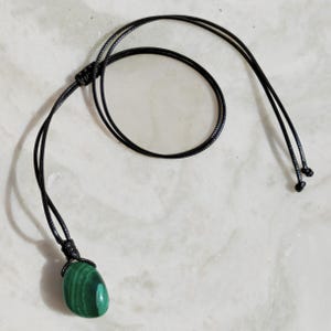 Close-up of a polished oval Malachite in shades of vivid green and deep forest green, resting on a light stone surface. The green opaque gemstone has a smooth glossy surface with distinct concentric banding, circular eye-like formations, and layered green gradients, with light reflections highlighting its rounded shape. It is attached to a black cord with a top knot and adjustable knot closure. Malachite is associated with transformation, protection, and emotional healing.