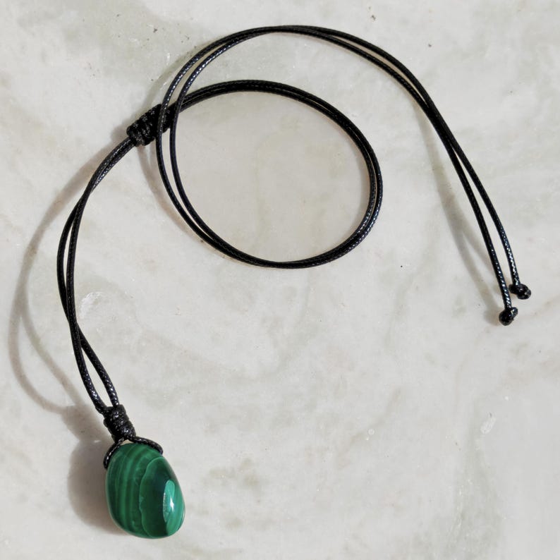 Close-up of a polished oval Malachite in shades of vivid green and deep forest green, resting on a light stone surface. The green opaque gemstone has a smooth glossy surface with distinct concentric banding, circular eye-like formations, and layered green gradients, with light reflections highlighting its rounded shape. It is attached to a black cord with a top knot and adjustable knot closure. Malachite is associated with transformation, protection, and emotional healing.