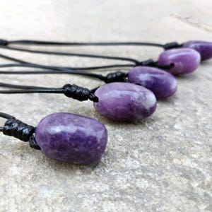 Five smooth, polished, rounded Lepidolite necklace pendants are displayed on a textured gray concrete surface. The purple gemstones vary slightly in color intensity and show some lighter patches and subtle striations. Each pendant is attached to a black cord. Lepidolite is associated with self-love, anti-anxiety, stress relief, transformation, emotional healing, and negativity release.