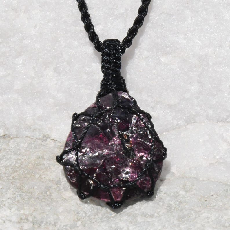 A single, raw, dtop shaped raw Garnet necklace pendant is displayed on a textured white rock surface. The dark reddish-purple gemstone has a rough, unpolished texture. It is encased in a black macrame net that forms the necklace cord. Garnet is associated with passion, energy, protection, and healing.