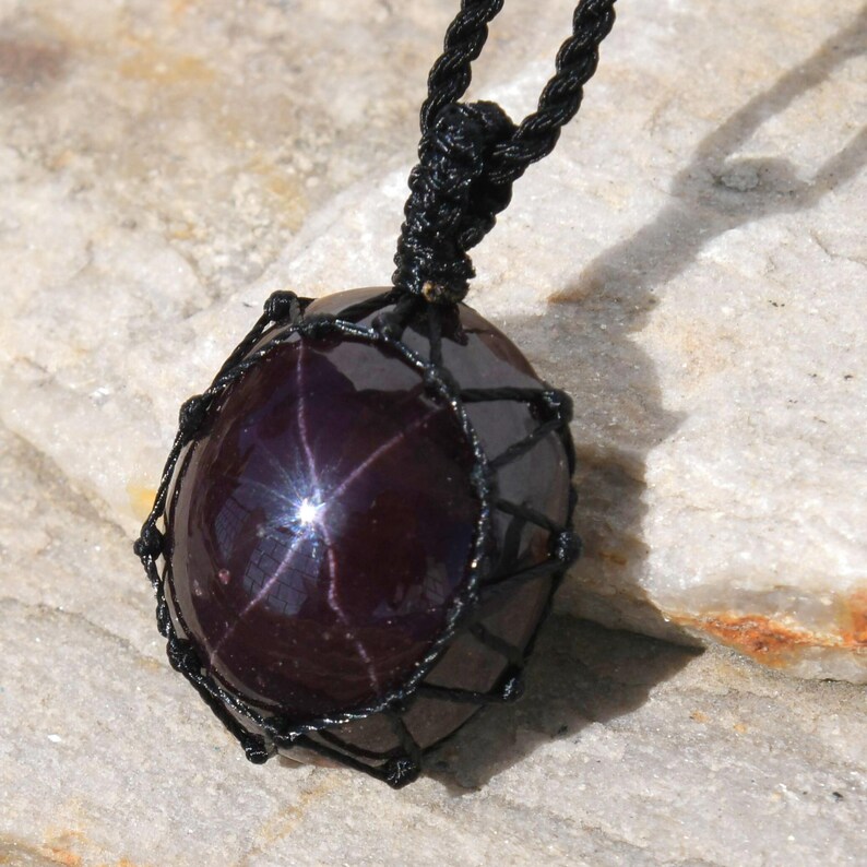 Close-up of a polished star garnet pendant in deep burgundy, wine-red, and dark plum tones, displayed on a light stone background. A distinct four-rayed star appears across the gemstone when illuminated, creating a bright reflective cross. The garnet has a smooth glossy finish with rich color depth and natural translucency. It is secured in a black macrame net and attached to a black adjustable cord. Associated with passion, vitality, grounding, courage, and emotional strength.