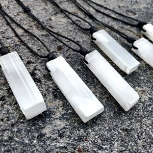 Five raw, rectangular selenite pendants hang from black cord necklaces, arranged diagonally on a textured gray stone surface with dark speckles. The white crystals have a striated, unpolished texture. These selenite necklaces are associated with cleansing and positive energy.