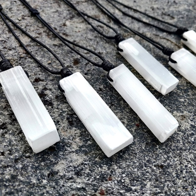Five raw, rectangular selenite pendants hang from black cord necklaces, arranged diagonally on a textured gray stone surface with dark speckles. The white crystals have a striated, unpolished texture. These selenite necklaces are associated with cleansing and positive energy.