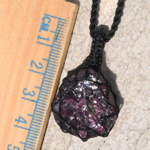Raw garnet crystal pendant in rich burgundy, raspberry-purple, and dark wine-red tones, displayed beside a wooden ruler on a light stone background. The natural gemstone features sparkling crystal faces, and vivid internal flashes that catch the light. Its rough organic texture highlights the crystal's natural formation. The pendant is secured in a black macrame net and attached to a black adjustable cord. Garnet is associated with passion, vitality, grounding, courage, and emotional strength.