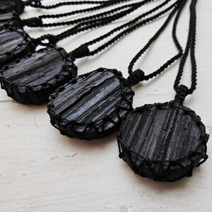 Close-up of multiple raw round Black Tourmaline in deep black shades, resting on a white wood surface. The Black Tourmaline dark gemstone has strong parallel vertical striations, rough uneven edges, and a matte to slightly reflective texture with natural ridges and occasional lighter inclusions and surface variations. Each piece is wrapped in a black macrame net and attached to a black cord with an adjustable knot closure. Black Tourmaline is associated with grounding and protection.