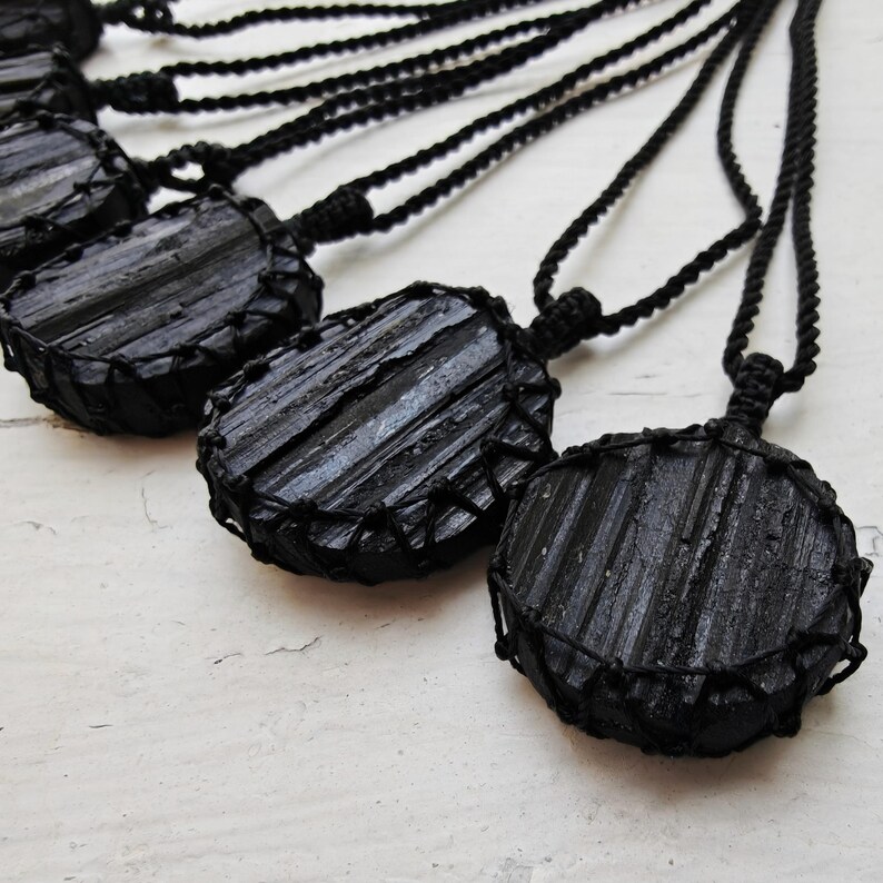 Close-up of multiple raw round Black Tourmaline in deep black shades, resting on a white wood surface. The Black Tourmaline dark gemstone has strong parallel vertical striations, rough uneven edges, and a matte to slightly reflective texture with natural ridges and occasional lighter inclusions and surface variations. Each piece is wrapped in a black macrame net and attached to a black cord with an adjustable knot closure. Black Tourmaline is associated with grounding and protection.