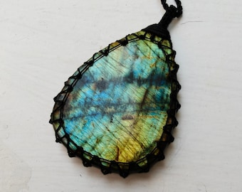 Puede incluir: Un colgante de labradorita en forma de lágrima con tonos azules y amarillos iridiscentes, encerrado en una red de cordón de macramé negro. La piedra muestra estrías naturales y una superficie pulida, suspendida de un collar de cordón negro.