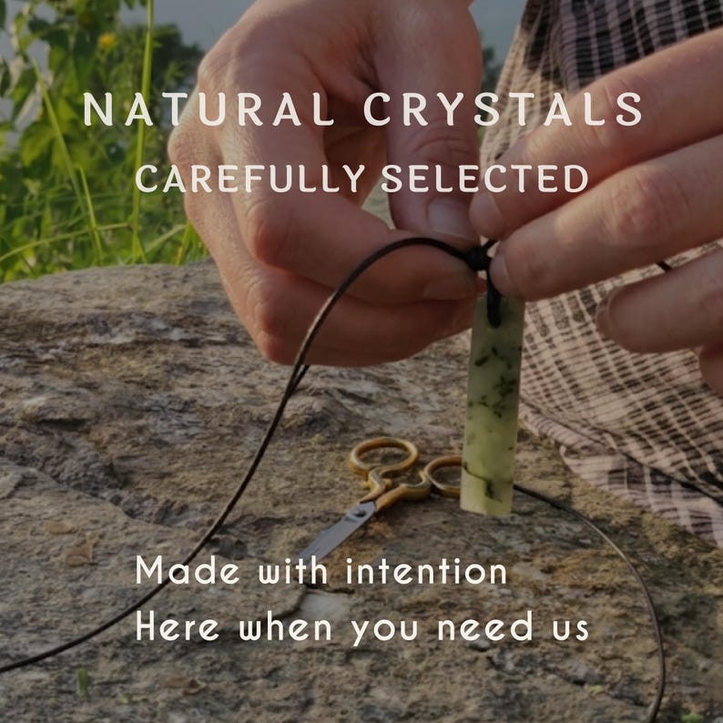 Close-up of hands crafting a gemstone necklace outdoors, threading a natural green crystal pendant onto a black cord. A pair of small scissors rests on the stone surface beside the work. Text on the image reads: "Natural Crystals Carefully Selected", "Made with intention", and "Here when you need us". The background features natural greenery and a rustic stone setting, emphasizing the handmade process and artisan craftsmanship.