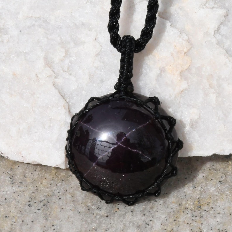 A single, smooth, polished, oval Star Garnet necklace pendant is displayed on a textured light rock surface. The dark red to reddish-purple gemstone exhibits a distinct four-rayed star (asterism) in its center when light reflects off it. It is encased in a black macrame net that forms the necklace cord. Star Garnet is associated with inner strength, protection, vitality, and regeneration.