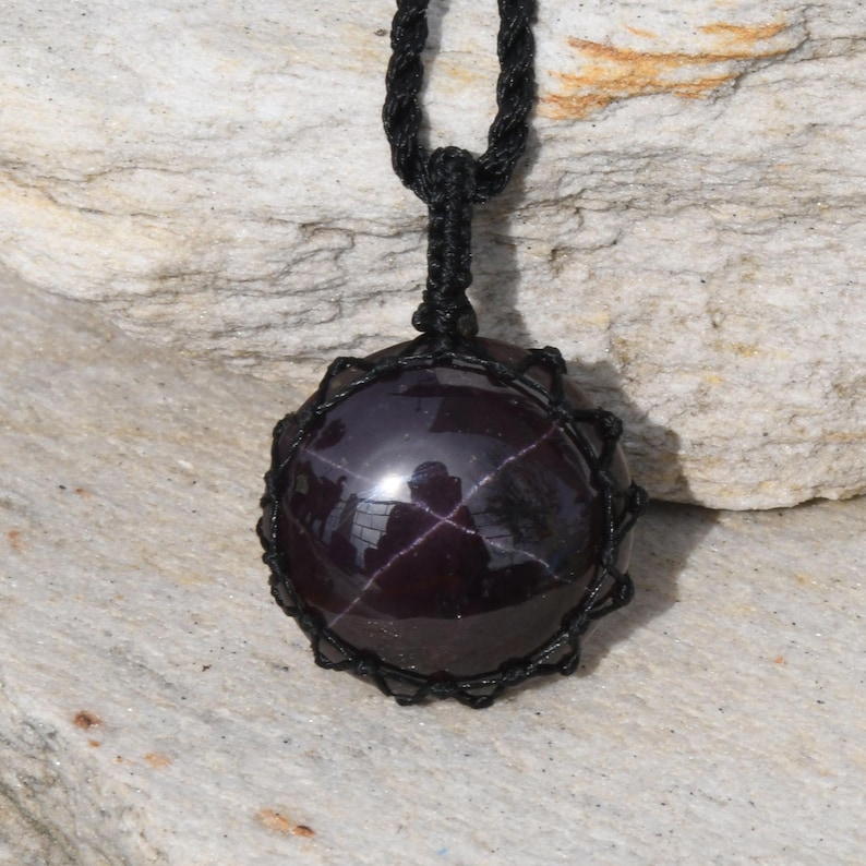 A single, smooth, polished, oval Star Garnet necklace pendant is displayed on a textured light rock surface. The dark red to reddish-purple gemstone exhibits a distinct four-rayed star (asterism) in its center when light reflects off it. It is encased in a black macrame net that forms the necklace cord. Star Garnet is associated with inner strength, protection, vitality, and regeneration.