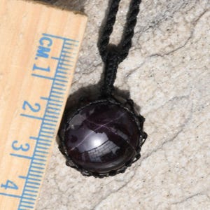 Close-up of one polished round Star Garnet in shades of deep burgundy and dark red, displayed on a light stone surface next to a ruler. The dark red Star Garnet gemstone has a smooth domed surface with a visible four-ray star reflection, glossy luster, and subtle internal inclusions. It is secured in a black macrame wrap and attached to a black cord with a top knot and adjustable knot closure. Star Garnet is associated with grounding, protection, and vitality.
