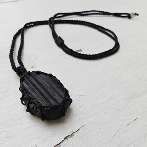 Close-up of 1 raw oval Black Tourmaline in deep black shades, resting on a white wood surface. The Black Tourmaline dark gemstone has strong parallel vertical striations, rough uneven edges, and a matte texture with natural ridges and subtle inclusions. It is wrapped in a black macrame net and attached to a black cord with an adjustable knot closure. Black Tourmaline is associated with grounding and protection.