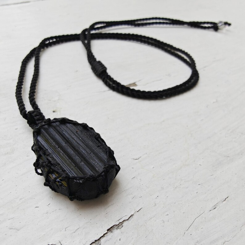 Close-up of 1 raw oval Black Tourmaline in deep black shades, resting on a white wood surface. The Black Tourmaline dark gemstone has strong parallel vertical striations, rough uneven edges, and a matte texture with natural ridges and subtle inclusions. It is wrapped in a black macrame net and attached to a black cord with an adjustable knot closure. Black Tourmaline is associated with grounding and protection.