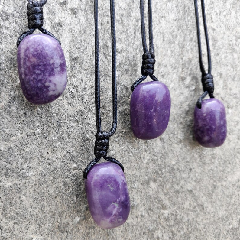 Four smooth, polished, rounded Lepidolite necklace pendants are displayed on a textured gray concrete surface. The purple gemstones vary slightly in color intensity and show some lighter patches and subtle striations. Each pendant is attached to a black cord. Lepidolite is associated with self-love, anti-anxiety, stress relief, transformation, emotional healing, and negativity release.