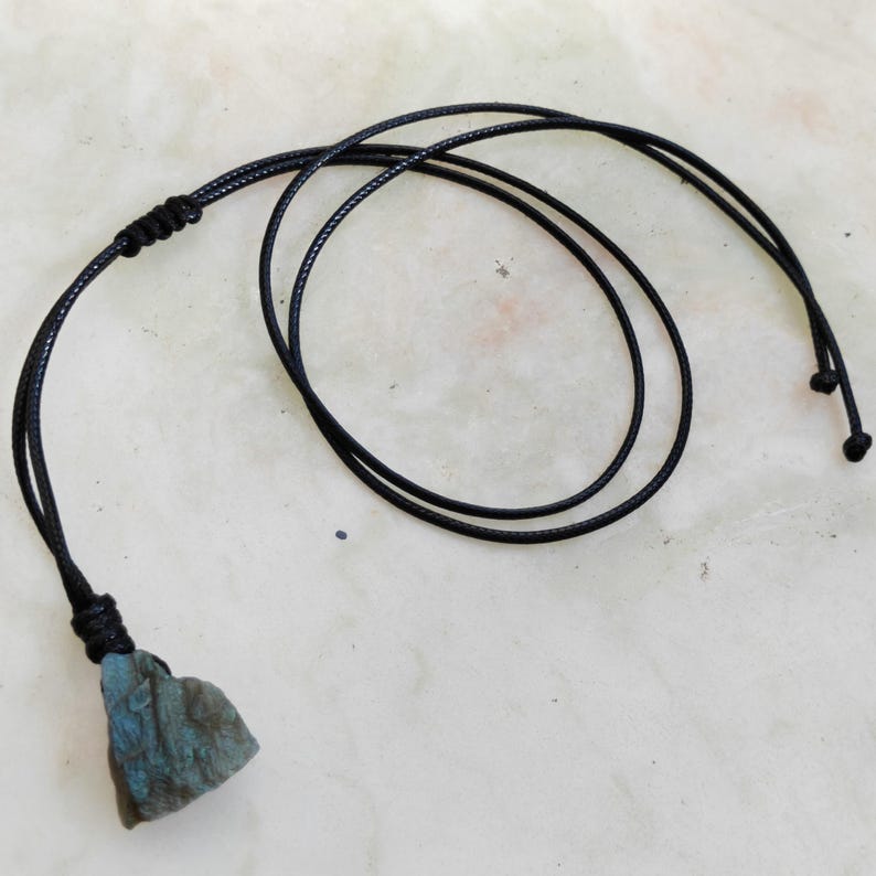 Close-up of a raw irregular Labradorite in shades of blue, and teal, resting on a light stone surface with the full black cord visible. The Labradorite gemstone has a natural rough texture with strong iridescent flashes, visible striations, and layered color play with reflective highlights. It is secured with a top knot and attached to a black cord with adjustable knot closure. Labradorite is associated with transformation, protection, intuition, and inner strength.