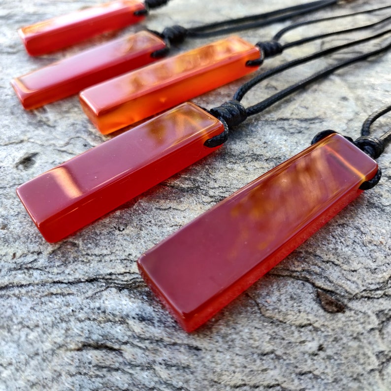 May include: Several rectangular pendants in shades of red and orange are displayed. Each pendant is suspended from a black cord. The pendants have a glossy, polished appearance. The background is a textured, gray surface.