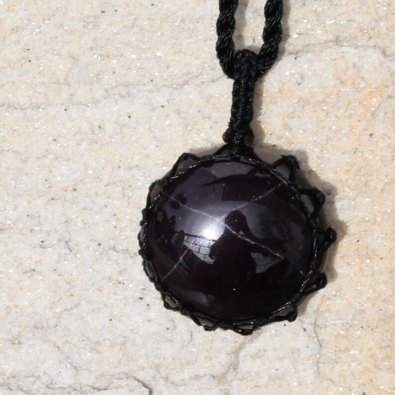 A single, smooth, polished, oval Star Garnet necklace pendant is displayed on a textured light rock surface. The dark red to reddish-purple gemstone exhibits a distinct four-rayed star (asterism) in its center when light reflects off it. It is encased in a black macrame net that forms the necklace cord. Star Garnet is associated with inner strength, protection, vitality, and regeneration.