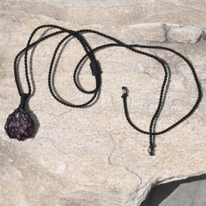 A single, raw, dtop shaped raw Garnet necklace pendant is displayed on a textured white rock surface. The dark reddish-purple gemstone has a rough, unpolished texture. It is encased in a black macrame net that forms the necklace cord. Garnet is associated with passion, energy, protection, and healing.