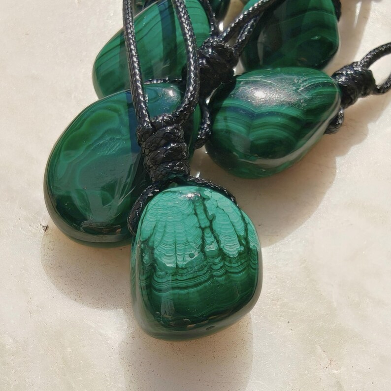 Close-up of multiple polished irregular Malachite in shades of vivid green and deep forest green, arranged on a light stone surface. The green opaque gemstones have smooth glossy surfaces with distinct banded patterns, concentric lines, and varied formations, including rounded and triangular shapes with layered green stripes and darker swirls. It is attached to a black cord with a top knot and adjustable knot closure. Malachite is associated with transformation, protection, and emotional healing
