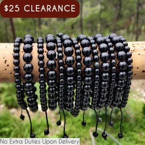 May include: Multiple strands of black beaded bracelets are displayed on a wooden dowel. The beads vary in size and are strung on black cords. The bracelets are against a blurred green background. The text "$25 CLEARANCE" is at the top.