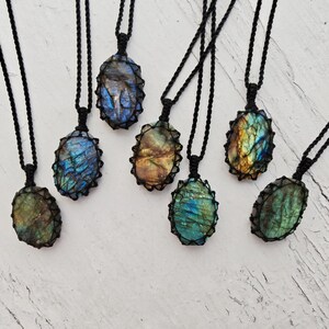 May include: Six oval-shaped labradorite pendants, each encased in a black macrame cord setting. The gemstones display iridescent flashes of blue, gold, and green. Each pendant hangs from a matching black braided cord necklace of equal length.