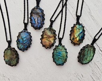 Puede incluir: Seis colgantes de labradorita de forma ovalada, cada uno engastado en un marco de cordón de macramé negro. Las piedras preciosas muestran destellos iridiscentes en azul, dorado y verde. Cada colgante cuelga de un collar de cordón trenzado negro.