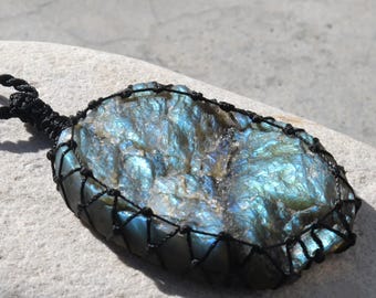Puede incluir: Colgante de labradorita iridiscente, mostrando una gama de azules y verdes. La piedra está engarzada en una red de macramé negra, con un cordón a juego. El colgante se exhibe sobre una superficie gris clara. Mide aproximadamente 7 cm de largo.