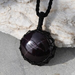 A single, smooth, polished, oval Star Garnet necklace pendant is displayed on a textured light rock surface. The dark red to reddish-purple gemstone exhibits a distinct four-rayed star (asterism) in its center when light reflects off it. It is encased in a black macrame net that forms the necklace cord. Star Garnet is associated with inner strength, protection, vitality, and regeneration.